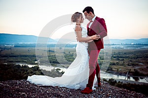 Cheerful newlywed couple is tenderly hugging on the mountains at the background of the landscape during the sunset.