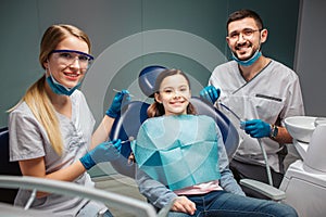 Cheerful nice young man woman detists sit and pose with child in dental chair. Adults hold tools. Sit in room together. photo