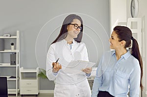 Cheerful nurse or doctor with clipboard talking to patient during consultation at clinic
