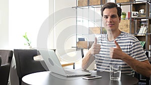 Cheerful office worker at desk doing thumb up sign and smiling. Real workplace