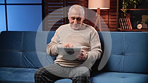 Cheerful old man using tablet for communicating with friends in social media, portrait in home