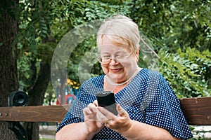 Cheerful old woman with smartphone at park. Outdoor portrait of happy senior woman at park using mobile phone. Plus size blond