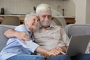 Cheerful older 60s grandparent couple using laptop on couch