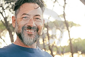 Cheerful outdoor portrait of happy adult mature man smiling at the camera with trees and sunset light in background. Beard and