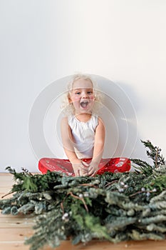 Cheerful overjoyed blonde female child sits crossed legs and Christmas garland in foreground, isolated over pink photo