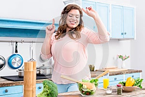 cheerful overweight woman listening music in headphones and dancing at table with fresh vegetables in kitchen