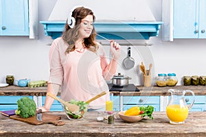 cheerful overweight woman listening music in headphones at table with fresh vegetables in kitchen