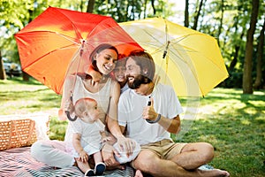 Cheerful parents with two kids have a rest on the lawn under the bright red and yellow umbrellas covering them from the