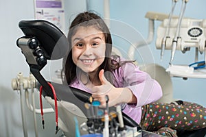 Cheerful patient girl showing thumbs up at dental clinic office. Medicine, stomatology and health care concept