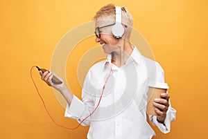 Cheerful pensioner woman with short haircut listens to music and holds a glass of coffee, dances on a yellow background