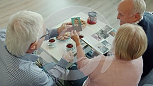 Cheerful pensioners watching pictures from photobook chatting relaxing at table with food