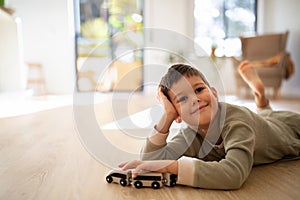 Cheerful pensive small european boy in pajamas lies on floor, plays with cars alone, enjoy rest and spare time