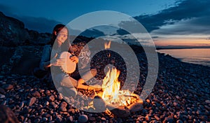 Cheerful percussionist girl playing djembe sitting on beach by the fire at sunset