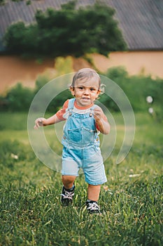 Cheerful perky toddler boy runs to meeting to camera with smile and happiness. Baby boy in denim overall in green park