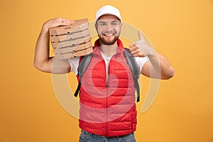 Cheerful pizza delivery man stands with carton boxes, waits for client, wears white cap and white tshirt with red waistcoat, poses