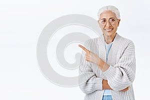 Cheerful pleasant senior woman, grandmother with grey combed hair, glasses, pointing upper left corner to promote