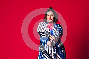 Cheerful plus size model in blue dress with big lollipop, happy fat woman on red background