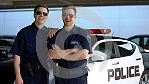 Cheerful police mates in sunglasses smiling into camera against squad car, work