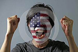 Cheerful portrait of a man with the flag of United States of America painted on his face on grey background.