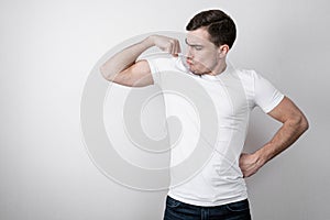 Cheerful portrait of young man in white blank t-shirt shows the bicep of his hand on gray background with copy space