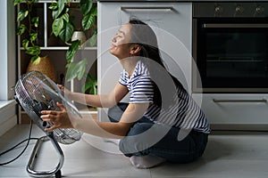 Cheerful positive Asian girl enjoys cold wind from electric fan sits on floor in kitchen of house