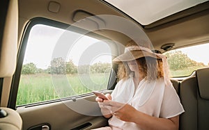 Cheerful positive curly young woman in casual wear with hat sitting in automobile backseat with fastened seatbelt and using phone