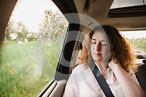 Cheerful positive curly young woman in casual wear sitting in automobile backseat with fastened seatbelt and looking away