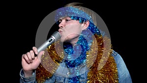 Cheerful, positive, handsome man holding microphone, listening to music and singing song with tinsel, isolated on black