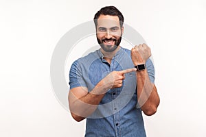 Cheerful positive man with beard in blue shirt pointing at wristwatch on hand and smiling, showing new smart clock, checking