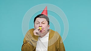 Cheerful positive middle aged man in party hat and white t-shirt with cardigan having fun blowing into party horn and thumbs up