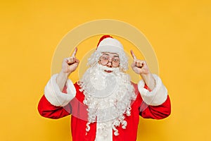 Cheerful and positive Santa Claus is pointing up on a copy space. Santa on a yellow background is raising his hands up,