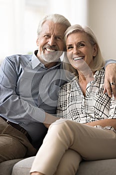 Cheerful positive senior retired husband and wife hugging on sofa