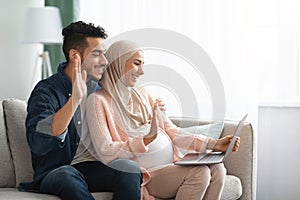 Cheerful Pregnant Muslim Couple Using Laptop At Home For Video Call