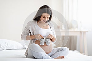 Cheerful pregnant woman holding baby shoes on big tummy