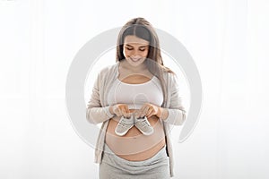 Cheerful pregnant woman holding baby shoes on big tummy