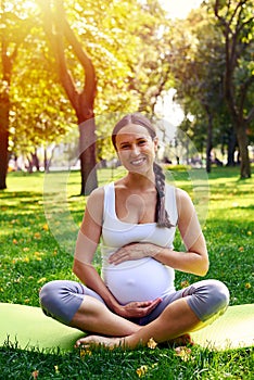 Cheerful pregnant woman holding belly on yoga mat in park