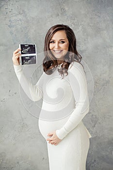 Cheerful pregnant woman showing ultrasound scan image and smiling on gray