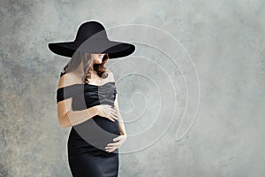 Cheerful pregnant woman in wide black broad brim hat portrait. Beautiful lady brunette wearing black dress ad smiling on grey