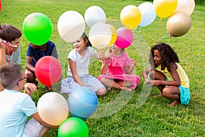 Cheerful preteen children sitting on lawn with balloons and talking