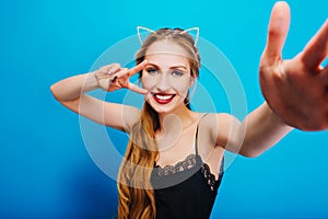 Cheerful pretty girl with cat ears in diamonds on head posing to camera, taking selfie, showing peace, enjoying party