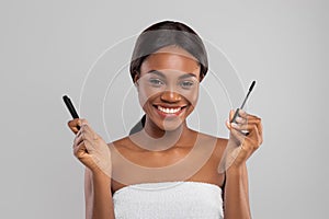 Cheerful pretty millennial african american female in towel show mascara for eyelashes and brush