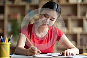 Cheerful pretty smart young chinese girl student making notes, homework, studying in living room