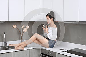 Cheerful pretty trendy stylish girl drinking coffee, sitting on table countertop in modern light white kitchen