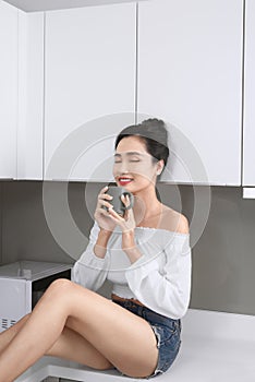 Cheerful pretty trendy stylish girl drinking coffee, sitting on table countertop in modern light white kitchen