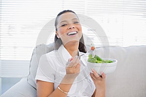 Cheerful pretty woman holding healthy salad sitting on sofa