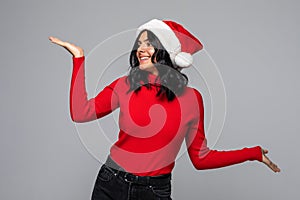 Cheerful pretty woman in santa hat making presentation of two products isolated on gray background