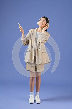 Cheerful pretty young asian woman listening to music with wireless headphones isolated over purple background, holding mobile