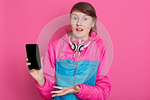 Cheerful pretty young Caucasian sportswoman holding blank screen smartphone and pointing on it isolated over pink background,
