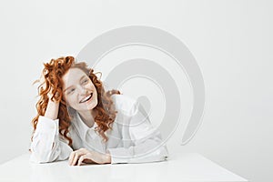 Cheerful pretty young girl with foxy hair smiling laughing sitting at table over white background.