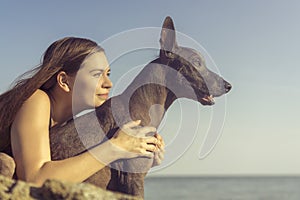 Cheerful pretty young girl sitting and hugging her dog xoloitzcuintli at the blue sky and sea on a stone beach